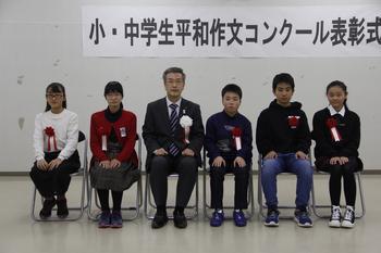胸に赤い胸章をつけた小学生の部受賞者5名とスーツを着た男性が横一列に並んで椅子に座っている写真