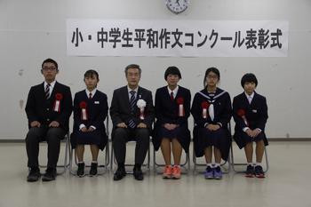 胸に赤い胸章をつけた中学生の部受賞者5名とスーツを着た男性が横一列に並んで椅子に座っている写真