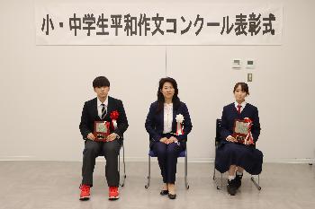 胸に赤い胸章をつけた中学生の部受賞者2名とスーツを着た女性が横一列に並んで椅子に座っている写真