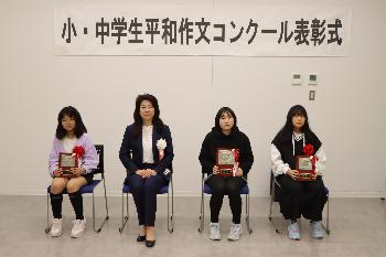 胸に赤い胸章をつけた小学生の部受賞者3名とスーツを着た女性が横一列に並んで椅子に座っている写真
