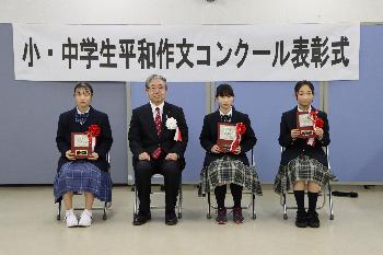 胸に赤い胸章をつけた中学生の部受賞者3名とスーツを着た男性が横一列に並んで椅子に座っている写真