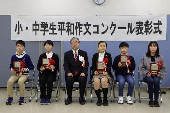 胸に赤い胸章をつけた小学生の部受賞者5名とスーツを着た男性が横一列に並んで椅子に座っている写真