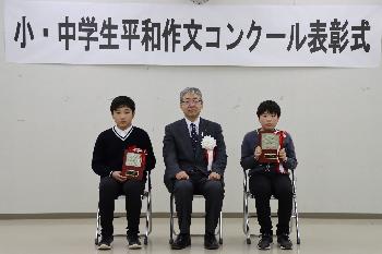 胸に赤い胸章をつけた小学生の部受賞者2名とスーツを着た男性が横一列に並んで椅子に座っている写真