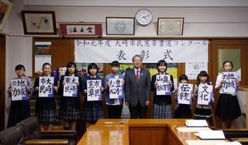「令和元年度大崎市民憲章書道コンクール表彰式」と書かれた横断幕の前で、で受賞者の小中学生が、書道の作品を持って、大崎市副市長と並んで写っている写真