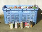 青色のボックスコンテナの中にたくさんのスチール缶が入っており、かごの前に色々な飲み物のアルミ缶が置かれている写真