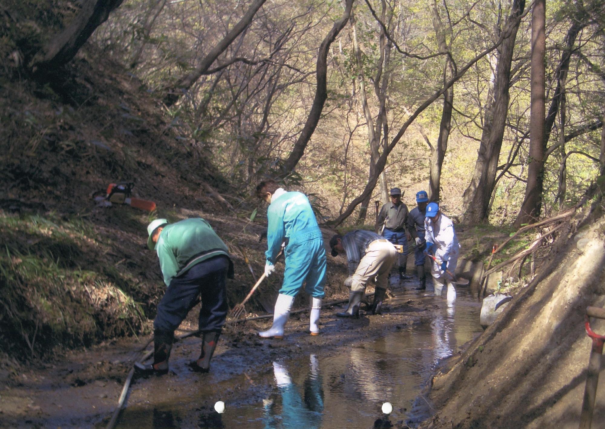 木々の生い茂った山道で長靴をはき、水管理の作業をしている5名の男性の写真