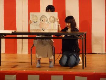 紅白の幕の前でゆきむすびの紙芝居をしている2人の女子大生の写真