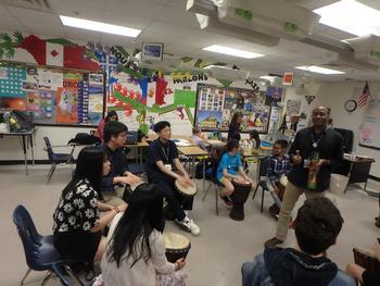 太鼓のような楽器を持った学生の皆さんが丸い円を作って椅子に座っており、中央に立っている先生が話をしてる音楽授業の様子の写真