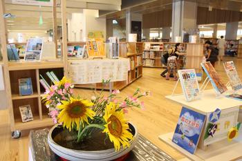 児童図書コーナー前に丸い花器に桃色の花と一緒にひまわりが生けられている生け花の写真