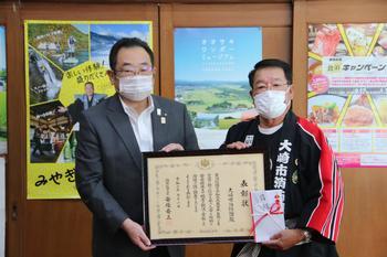 消防団の法被を着た大崎市消防団の男性と市長が、内閣総理大臣表彰で授与された表彰状と目録を持って、並んで写っている写真
