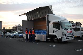 トラックの荷台にブルーシートが詰め込まれており、トラックの前にオレンジ色のベストを着た派遣職員3名が並んで立っている写真