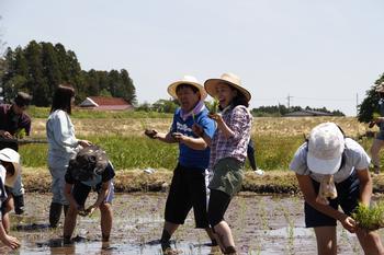 麦わら帽子を被ったワッキー貝山さんと佐藤千日さんが田んぼの中で片手に苗を持って笑顔で写っている写真