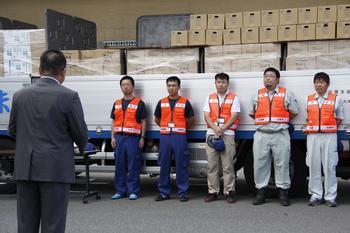 トラックの荷台にダンボール詰めされたペットボトルが積まれており、その前にオレンジ色のベストを着た派遣職員5名が整列して市長の話を聞いている様子の写真