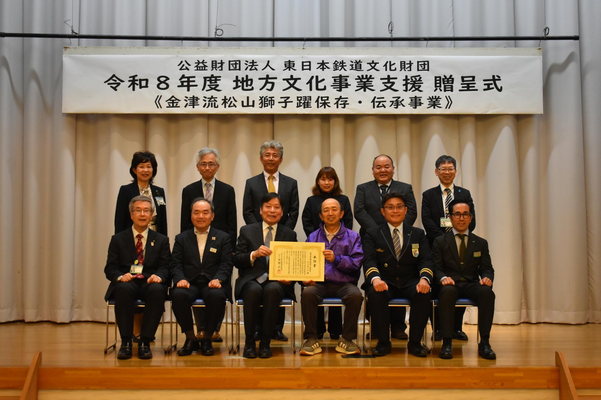 贈呈式の記念撮影。中央で日野理事長と及川会長が承認書を持ち、関係者がその周りに並んでいる。