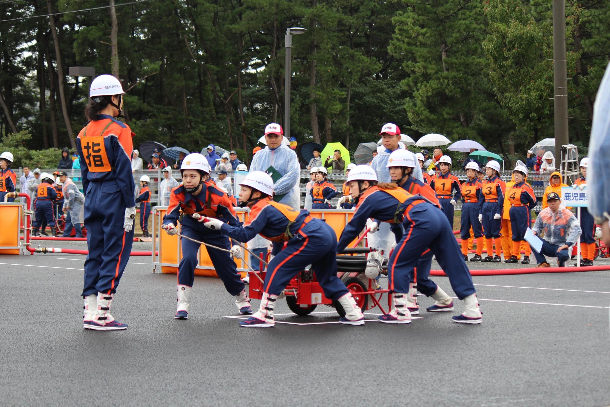 ヘルメットをかぶって活動服を着た女性消防団員4名がホースを乗せた赤い押し車を押して前方の2名がホースを持って「指」と書かれたゼッケンをつけた女性にホースを向けている訓練の様子の写真