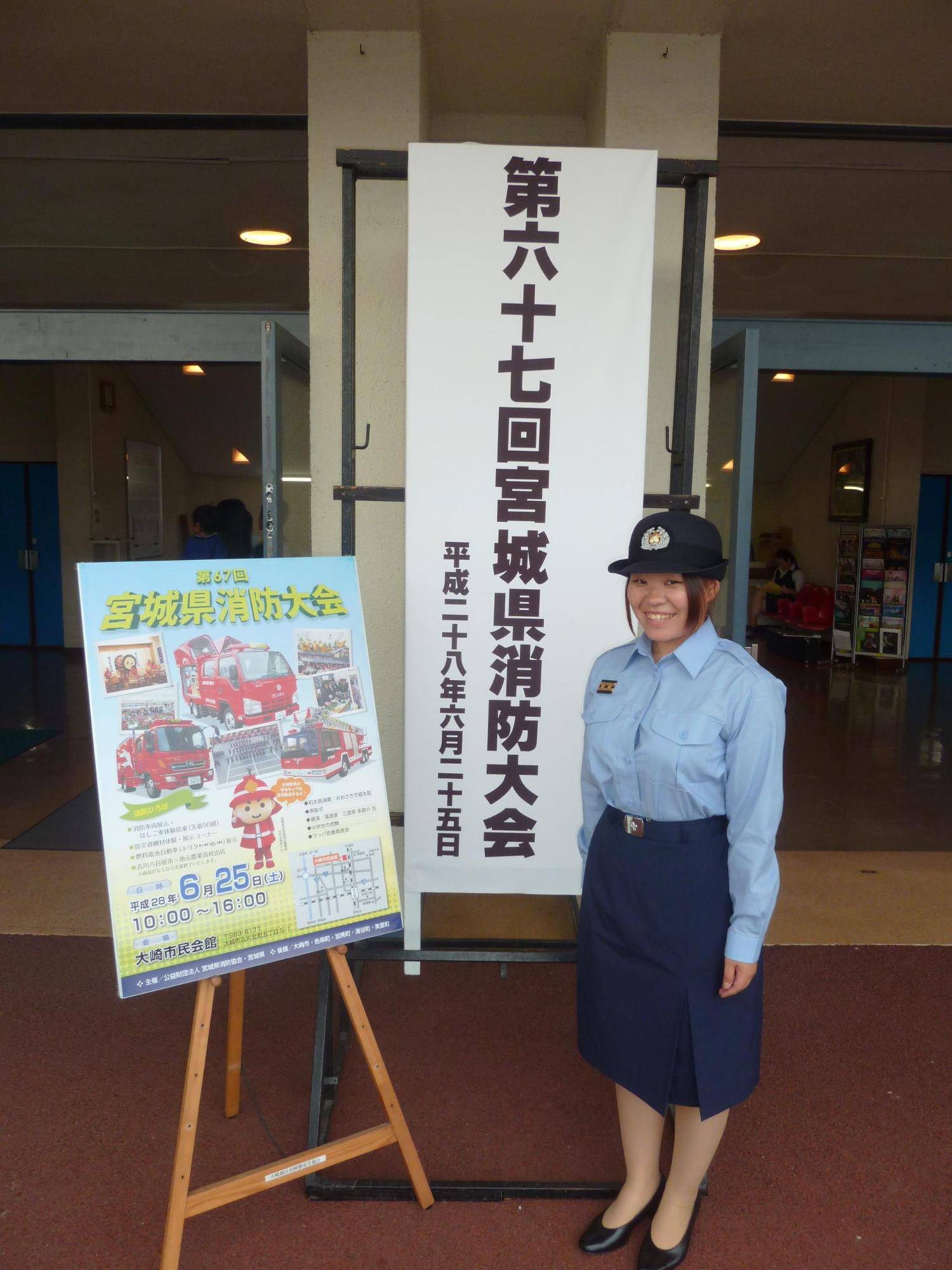 制服を着た女性消防団員が第六十七回宮城県消防大会と書かれた看板とポスターの前で笑顔で立っている写真